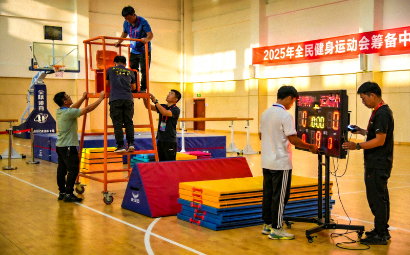 2025年全民健身运动会筹备 - 明亮体育馆内工作人员布置比赛场地与器材的画面